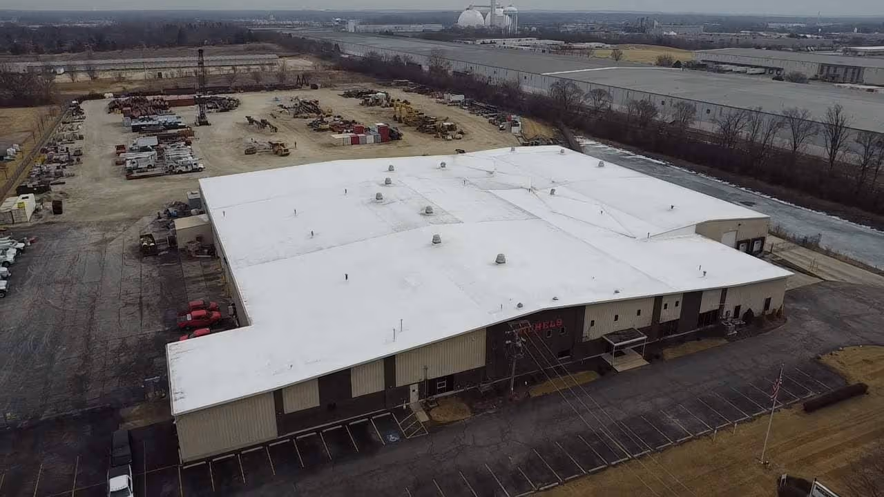 A large industrial warehouse with a white roof surrounded by parking lots and an adjacent dirt lot with construction equipment.
