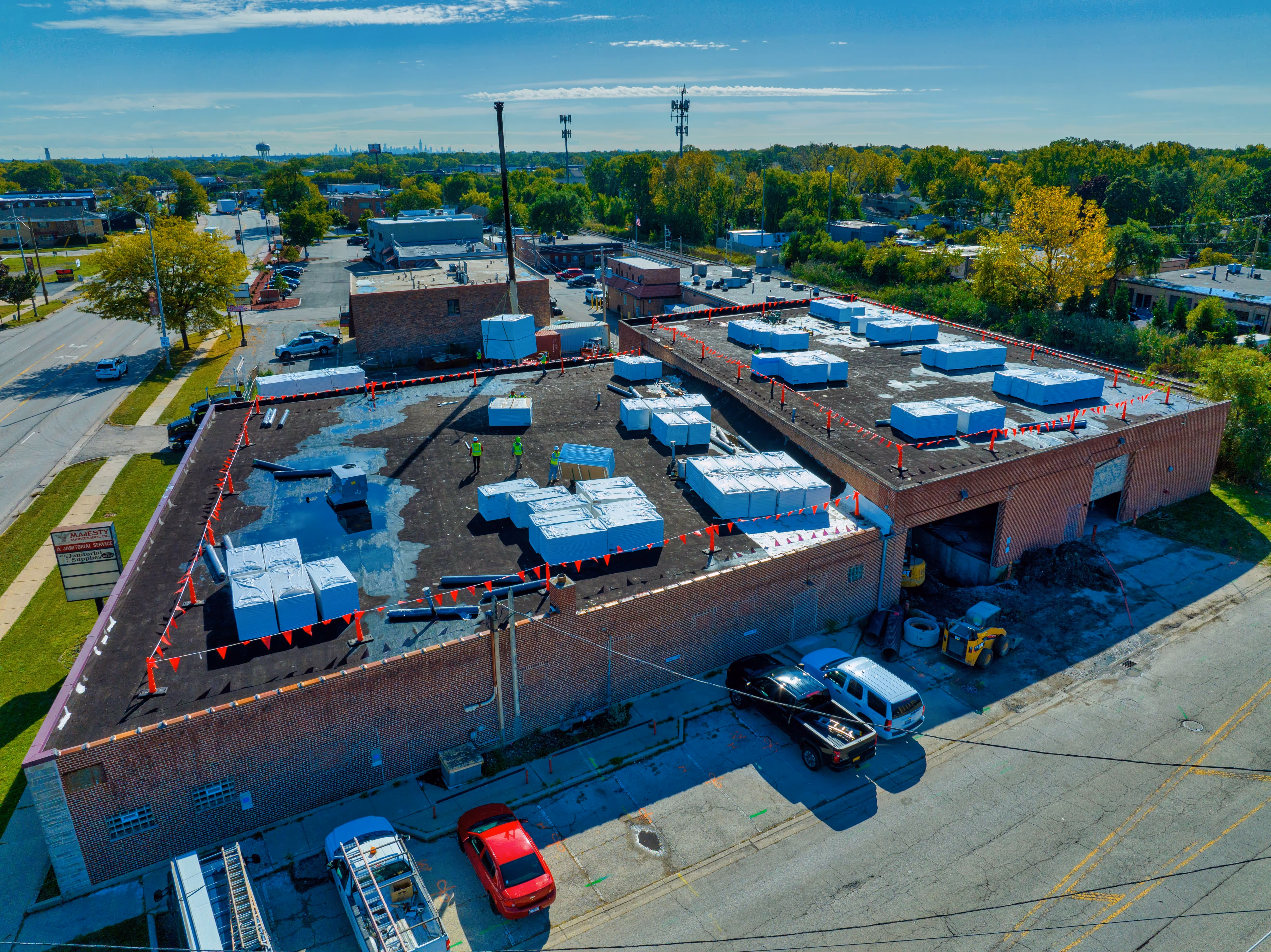 Aerial view of a large flat rooftop with stacks of construction materials and workers wearing safety vests, surrounded by red caution tape.