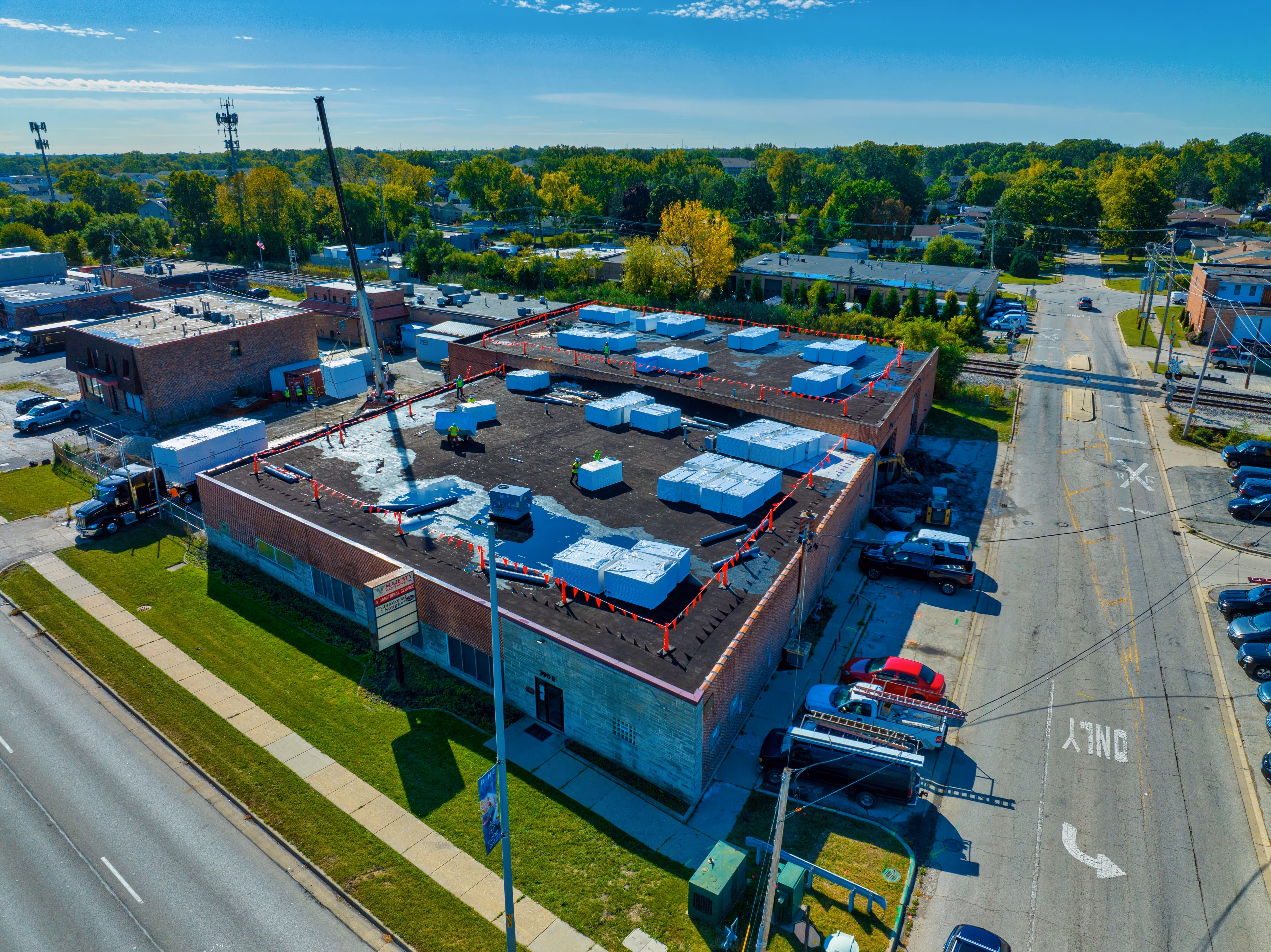 Aerial view of workers installing new insulation panels on the flat roof of a large brick commercial building surrounded by streets and parked vehicles.