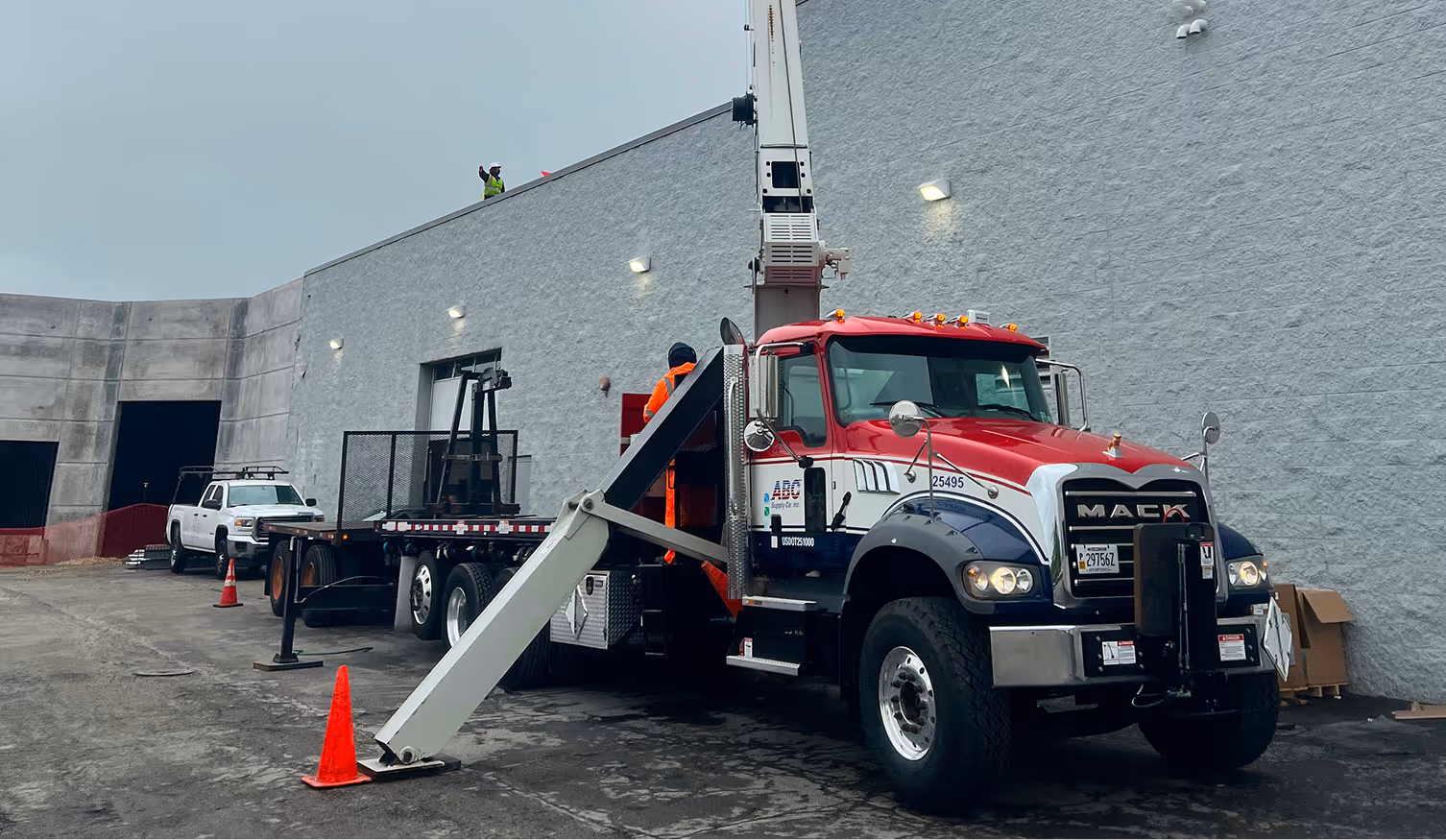 Red, white, and blue Mack crane truck parked next to a gray building with a worker in an orange safety suit operating the crane.