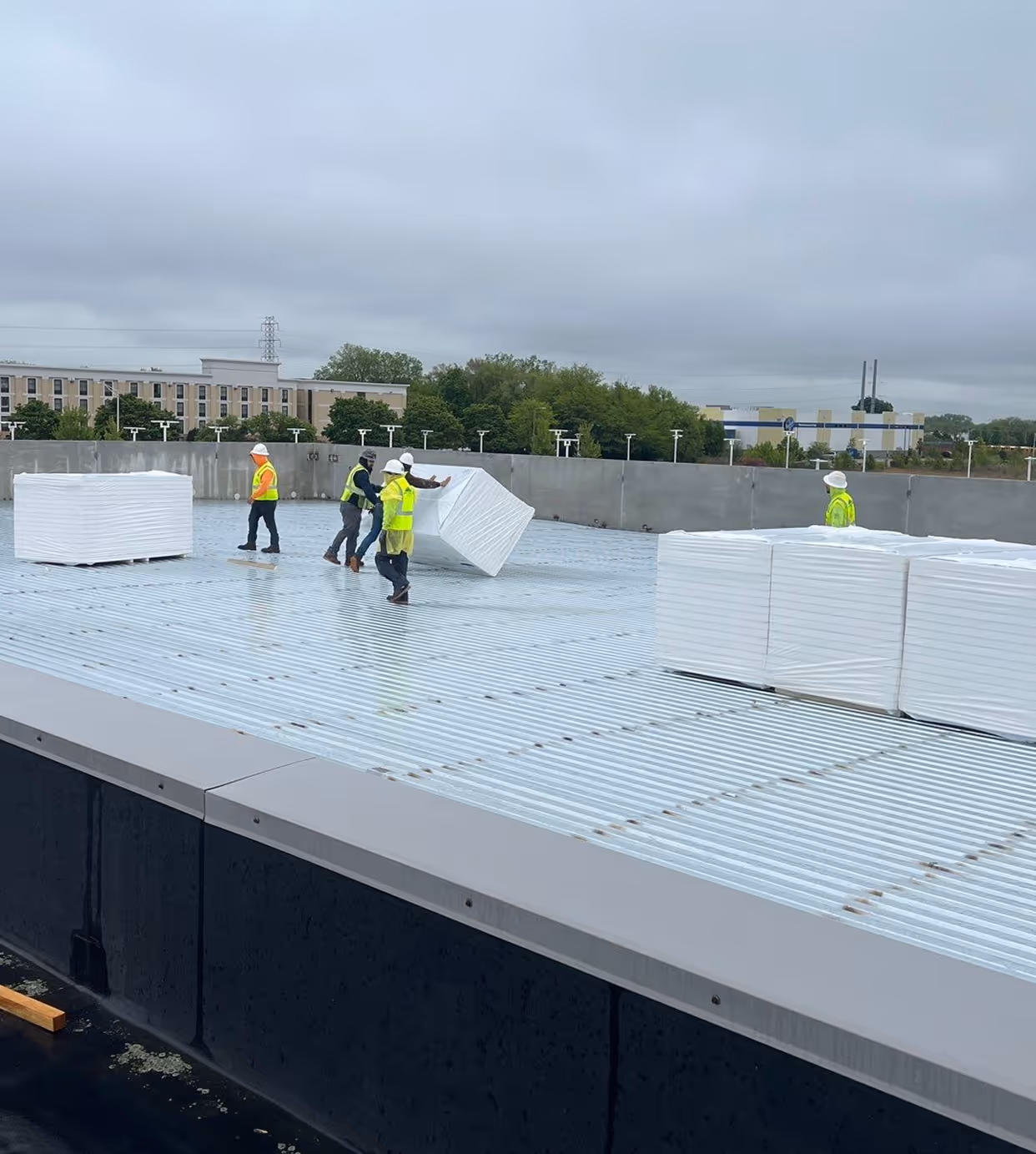 Construction workers in safety vests and helmets handling white insulation panels on a metal rooftop under a cloudy sky.