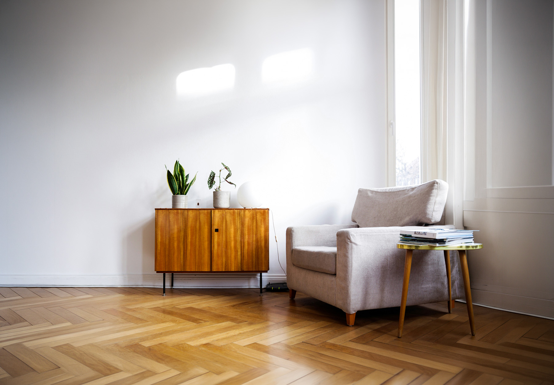 Bright living room corner with a small wooden cabinet holding two potted plants and a round white lamp, beside a beige armchair and wooden parquet floor.