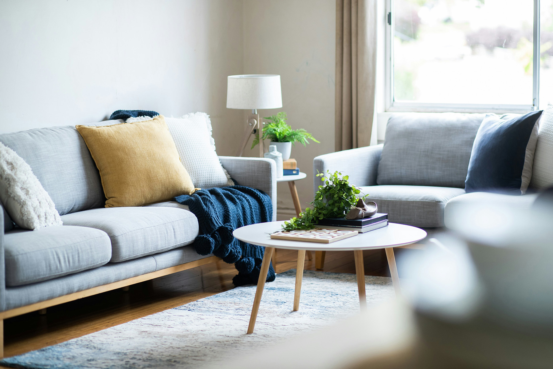 Bright living room with two light gray sofas, decorative pillows, a blue knitted throw, a round white coffee table with a plant and books, and a side table with a lamp and plant.