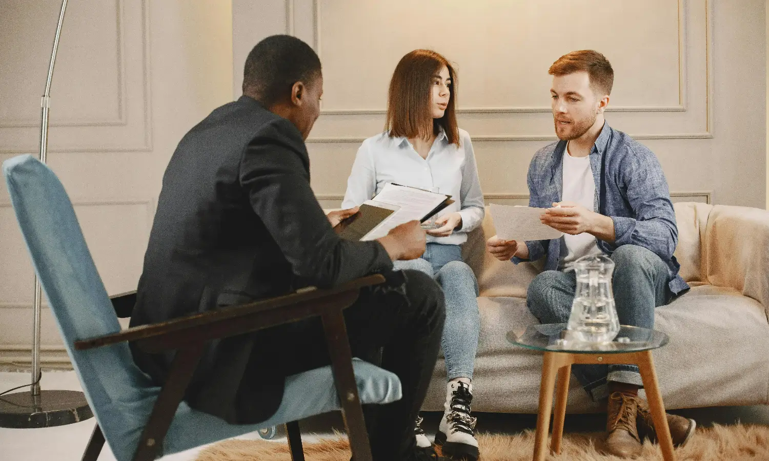 Three people having a serious discussion in a comfortable room, two sitting on a beige couch and one in a blue armchair holding papers and a tablet.