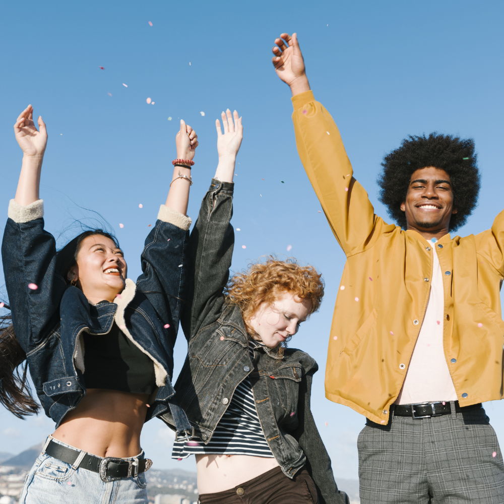 Three joyful young adults with raised arms celebrating outdoors under a clear blue sky.