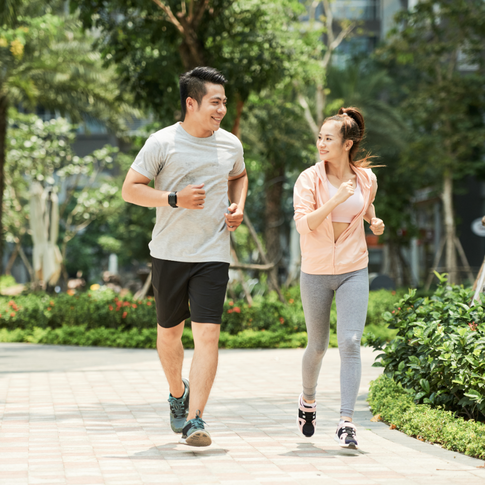 A smiling man and woman jogging together on a paved path in a park with lush green trees and bushes.