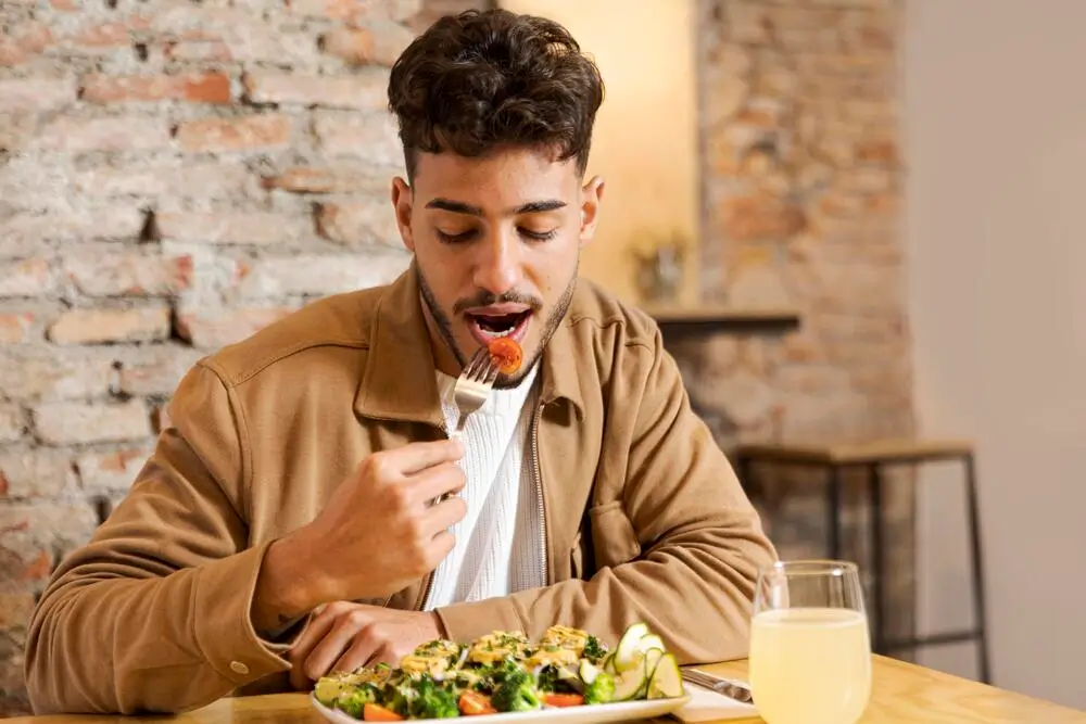 Man wearing a brown jacket eating a cherry tomato from a fork with a plate of grilled vegetables in front of him.