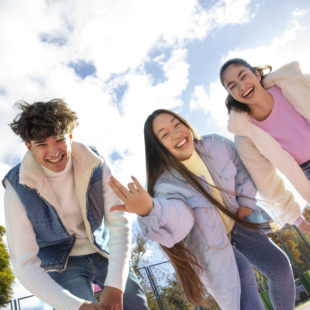 Three smiling teens outdoors on a sunny day, dressed in warm jackets against a blue sky with clouds.