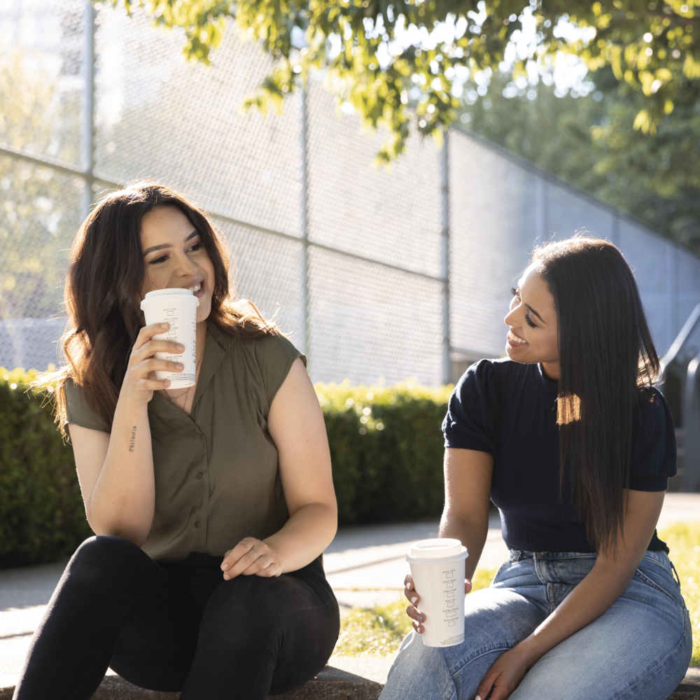 Two women sitting outside on a sunny day, smiling and holding takeaway coffee cups during a conversation.