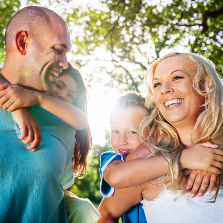 Smiling parents each carrying a child on their back outdoors in sunlight with greenery in the background.