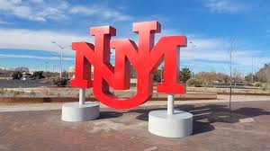 Large red UNM sculpture outdoors with a clear blue sky and paved ground.