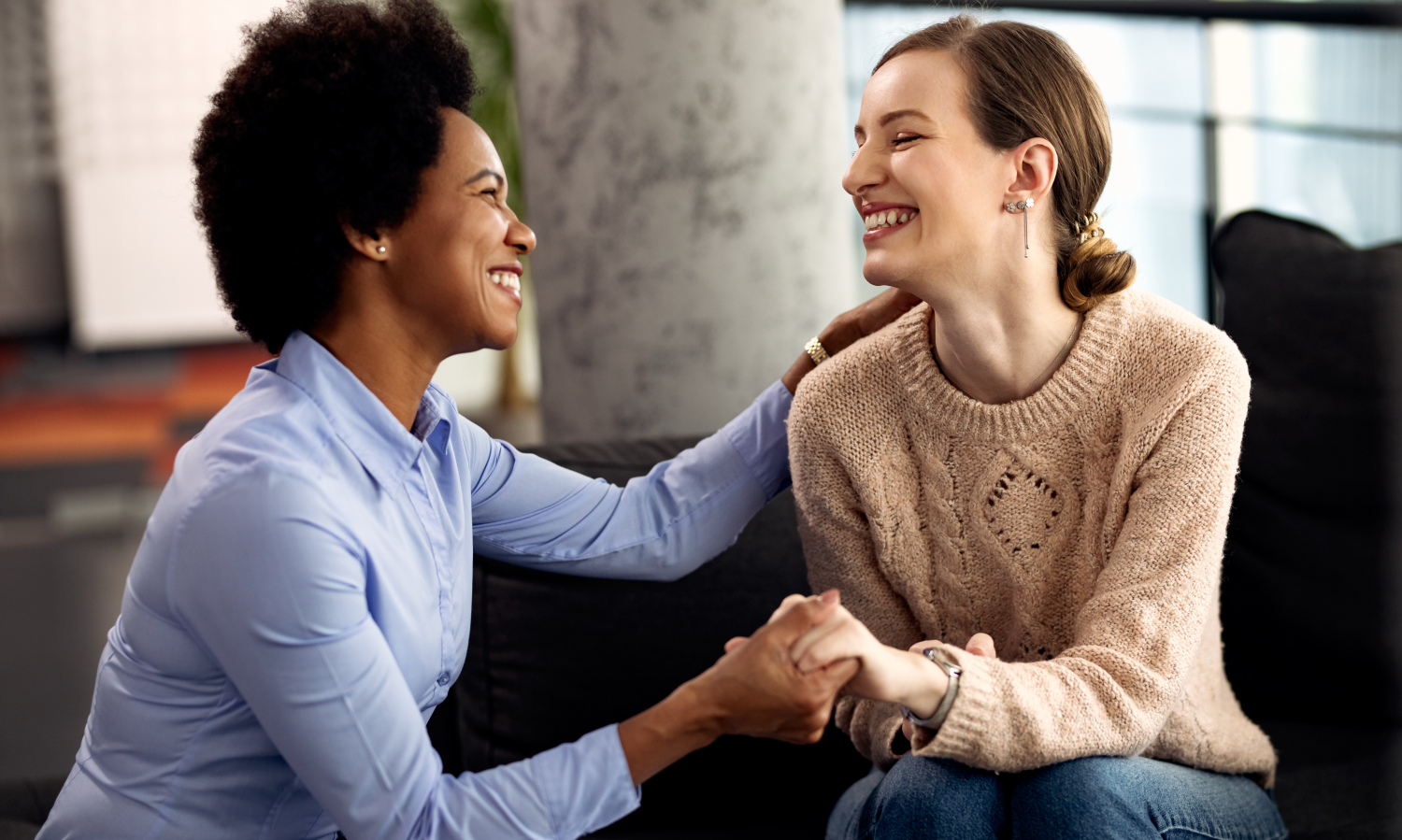 Two women sitting on a couch smiling warmly while holding hands and engaging in a supportive conversation.