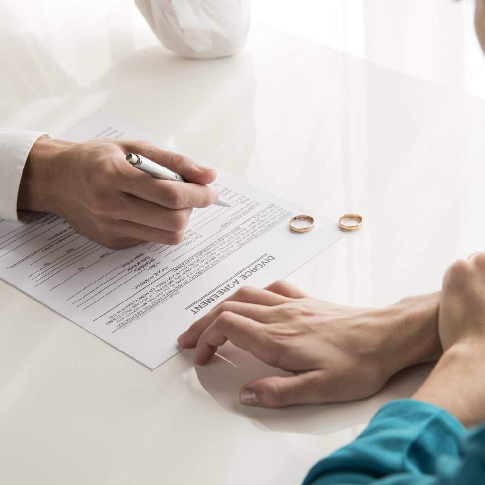 Person signing a divorce agreement on a desk with two wedding rings placed nearby.