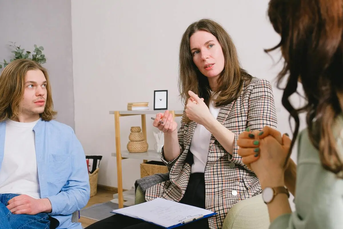 Woman therapist speaking and gesturing with a pen while two clients listen attentively in a cozy counseling session.