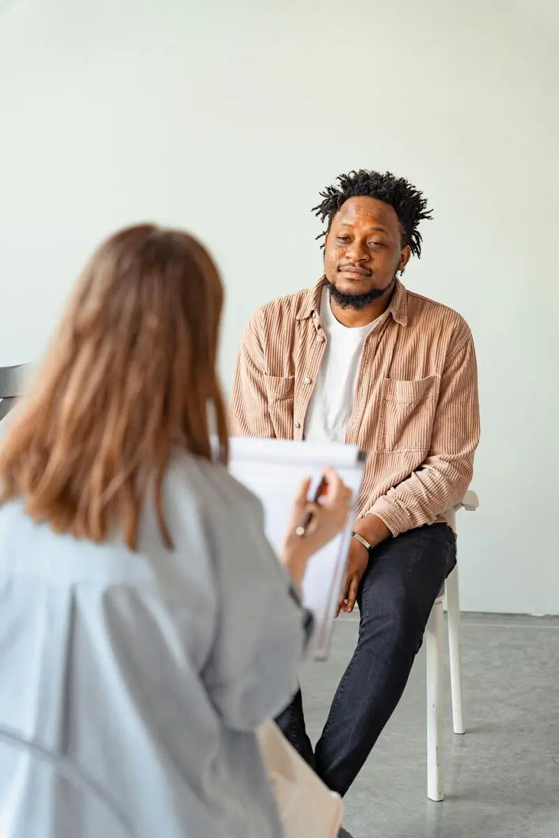 Man with a beard and afro hairstyle, wearing a brown jacket and white t-shirt, sitting on a chair facing a woman who is taking notes on a clipboard during a therapy session.