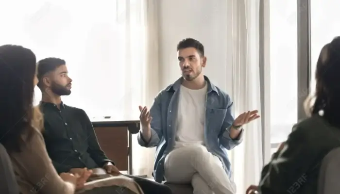 A man speaking gesturally in a group therapy session with three attentive listeners.