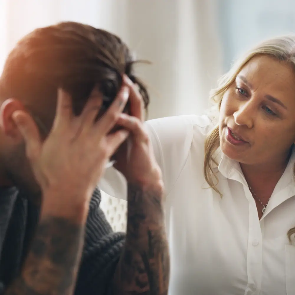 A healthcare provider in a white coat speaks with a distressed patient who is holding their head in their hands.