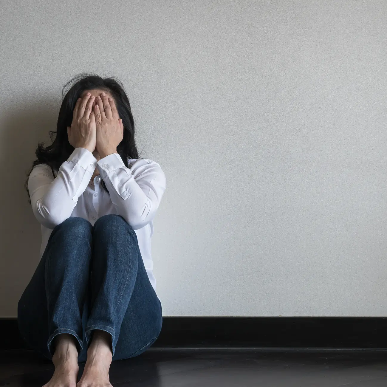 A woman sitting on the floor against a wall with her face buried in her hands.