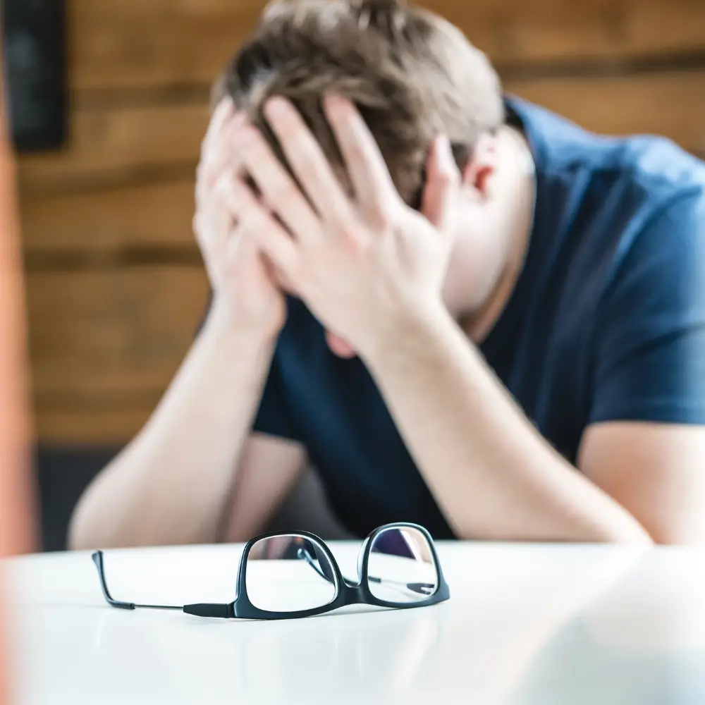 A man sitting at a desk with his head in his hands, his glasses set down in front of him.