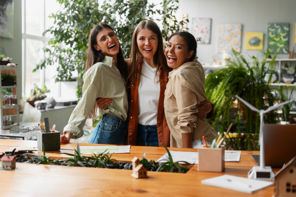 Three smiling women standing and hugging in a bright workspace with plants and drawings in the background.