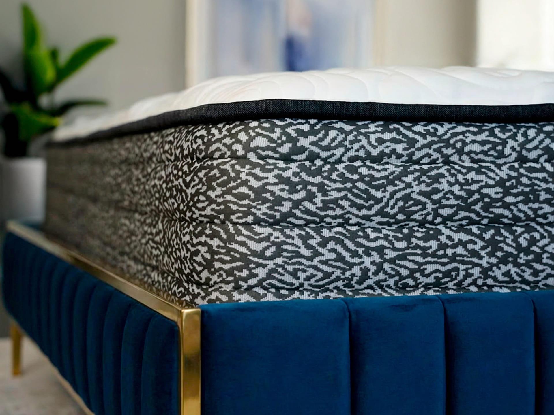 Close-up of a mattress with a white quilted top and black patterned side sitting on a blue upholstered bed frame with gold accents.