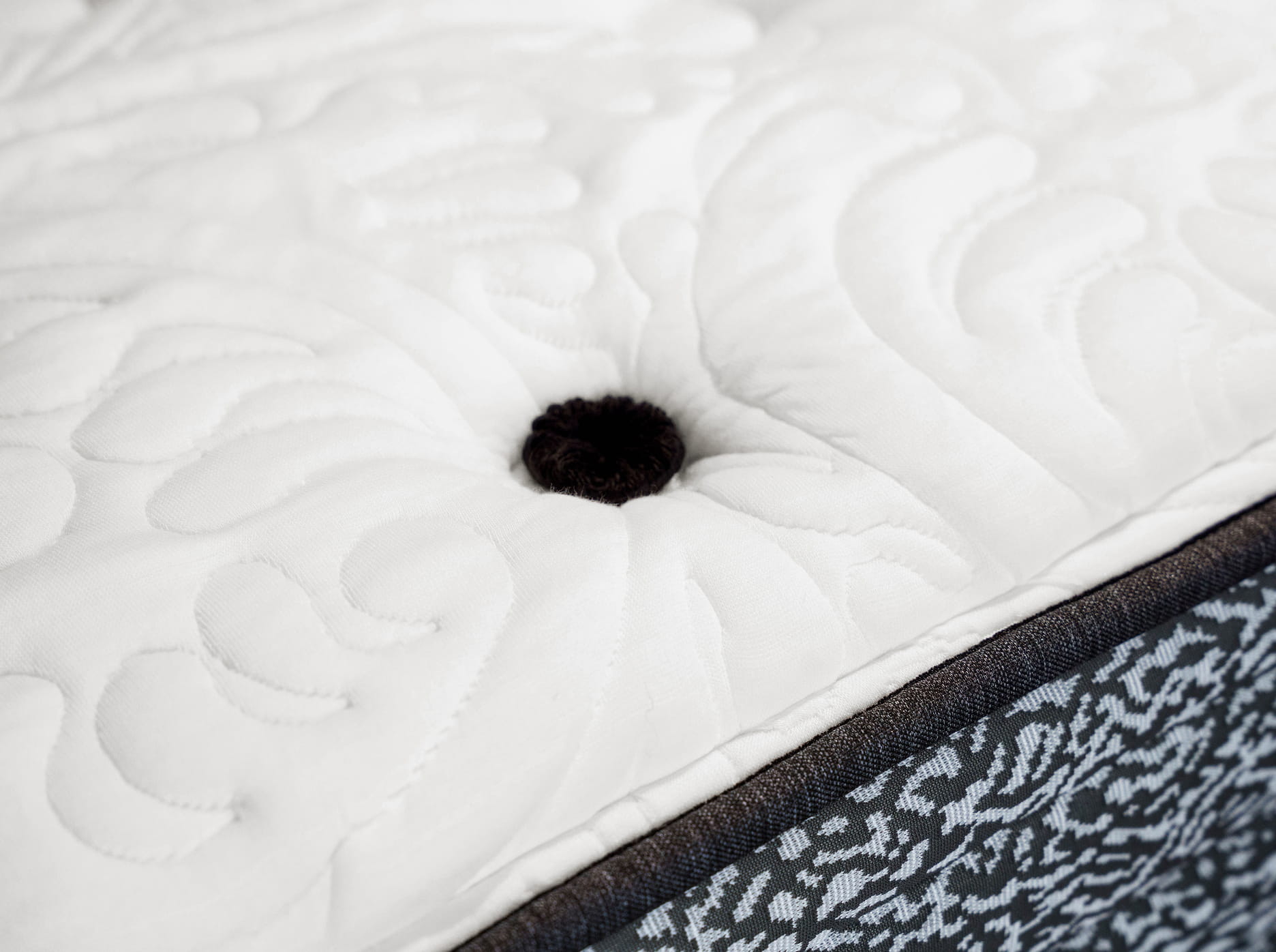 Close-up of a white quilted mattress top with a black button tuft and patterned side fabric.