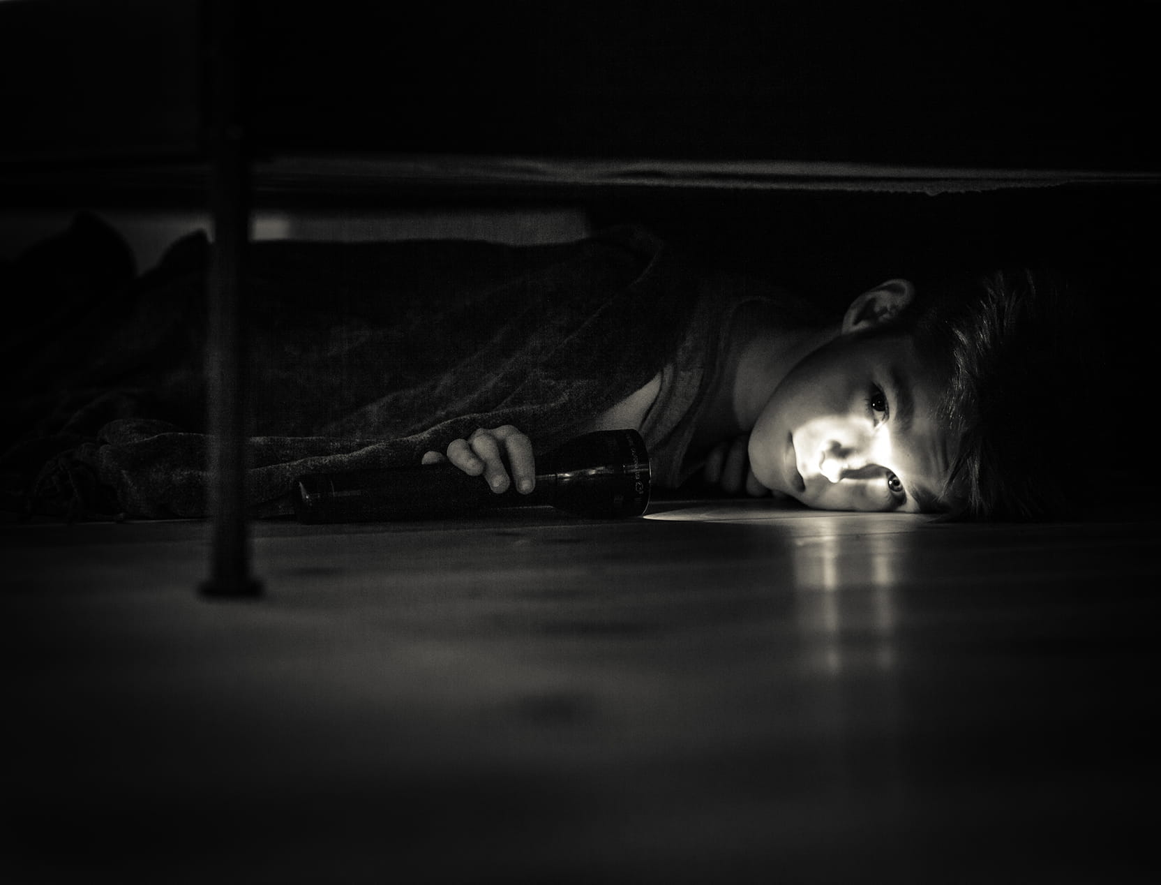 Boy lying on the floor under a bed, holding a flashlight that illuminates his face in a dark room.
