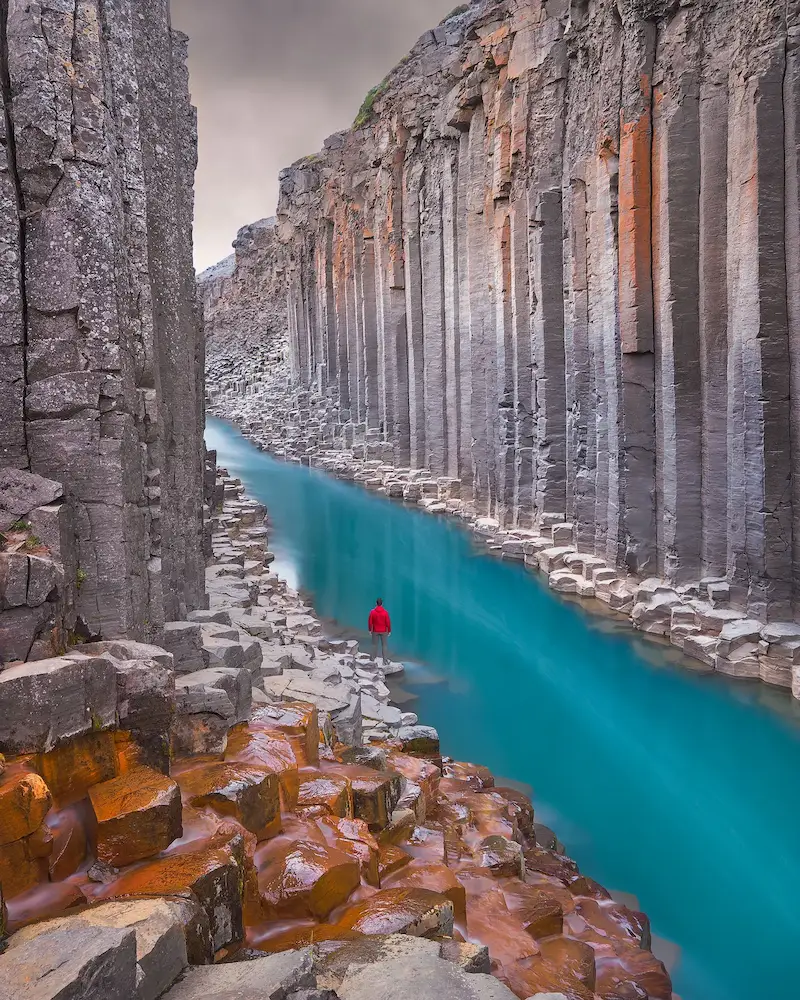 Cañón de columnas de basalto con un río turquesa y una figura de rojo, con las rocas anaranjadas del primer plano muy saturadas