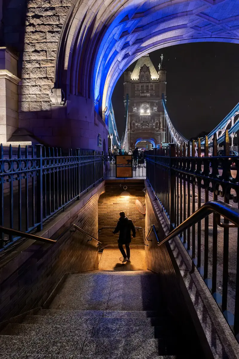 Escalera bajo el Tower Bridge de noche, con el arco iluminado de azul intenso compitiendo con la luz cálida de la escalera