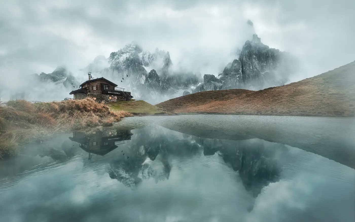 Refugio de montaña junto a un lago en calma, con las cumbres entre la niebla reflejadas en el agua y la línea de orilla cerca del centro del encuadre
