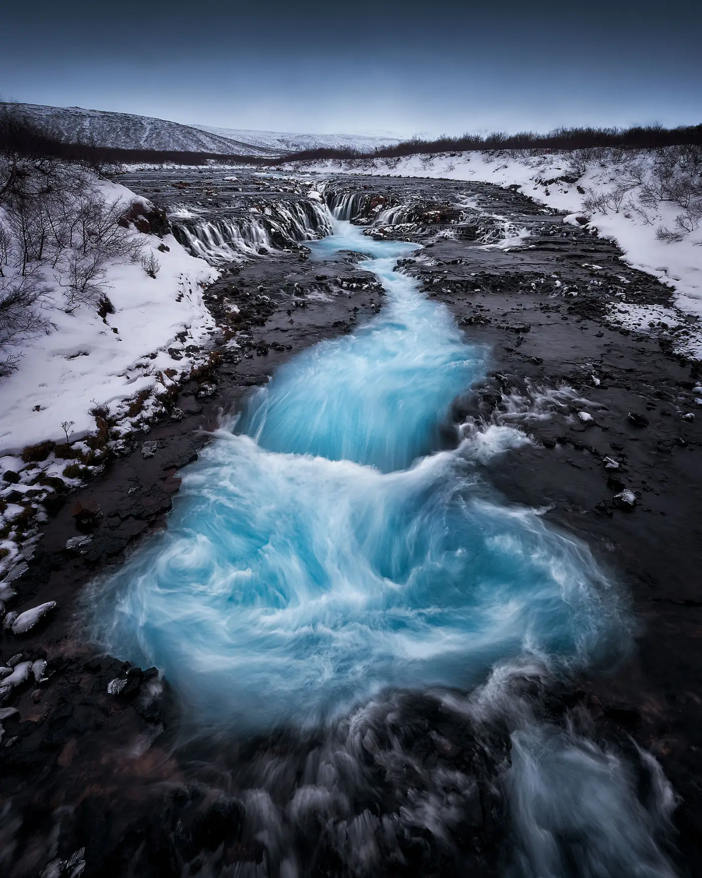 Cascada islandesa de agua turquesa fotografiada desde muy cerca del cauce, con el agua ocupando todo el primer plano y el paisaje nevado alejándose hacia el fondo