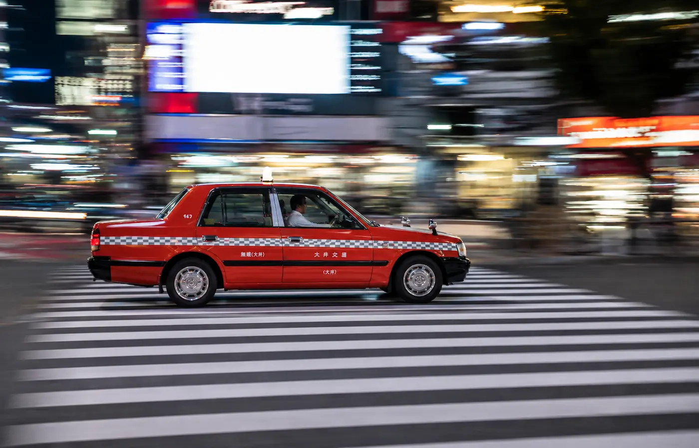 Taxi rojo cruzando un paso de peatones de noche: el coche sale definido mientras los edificios y los luminosos del fondo aparecen estirados por el movimiento de la cámara
