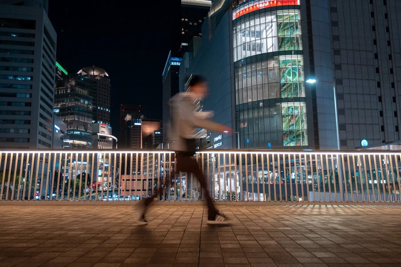 La misma persona cruzando la pasarela fotografiada a 1/15 de segundo: la figura se deshace en un rastro mientras la barandilla y los edificios siguen definidos
