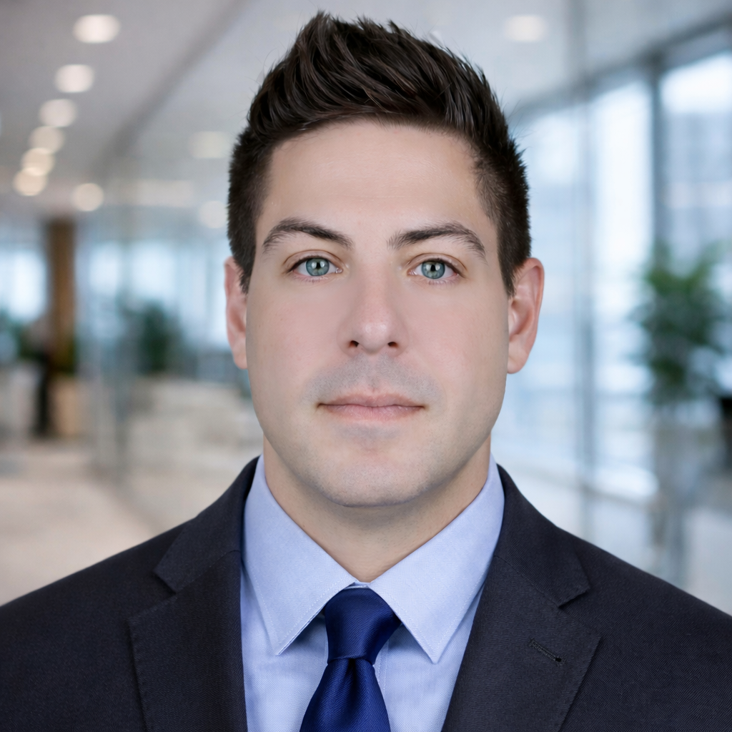 Professional man with dark hair wearing a dark suit, light blue shirt, and navy tie standing in a modern office.
