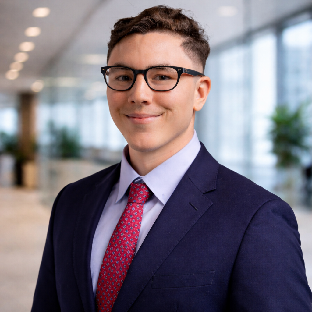 Smiling young man with short curly hair, black glasses, wearing a navy suit, light blue shirt, and red patterned tie in a modern office.