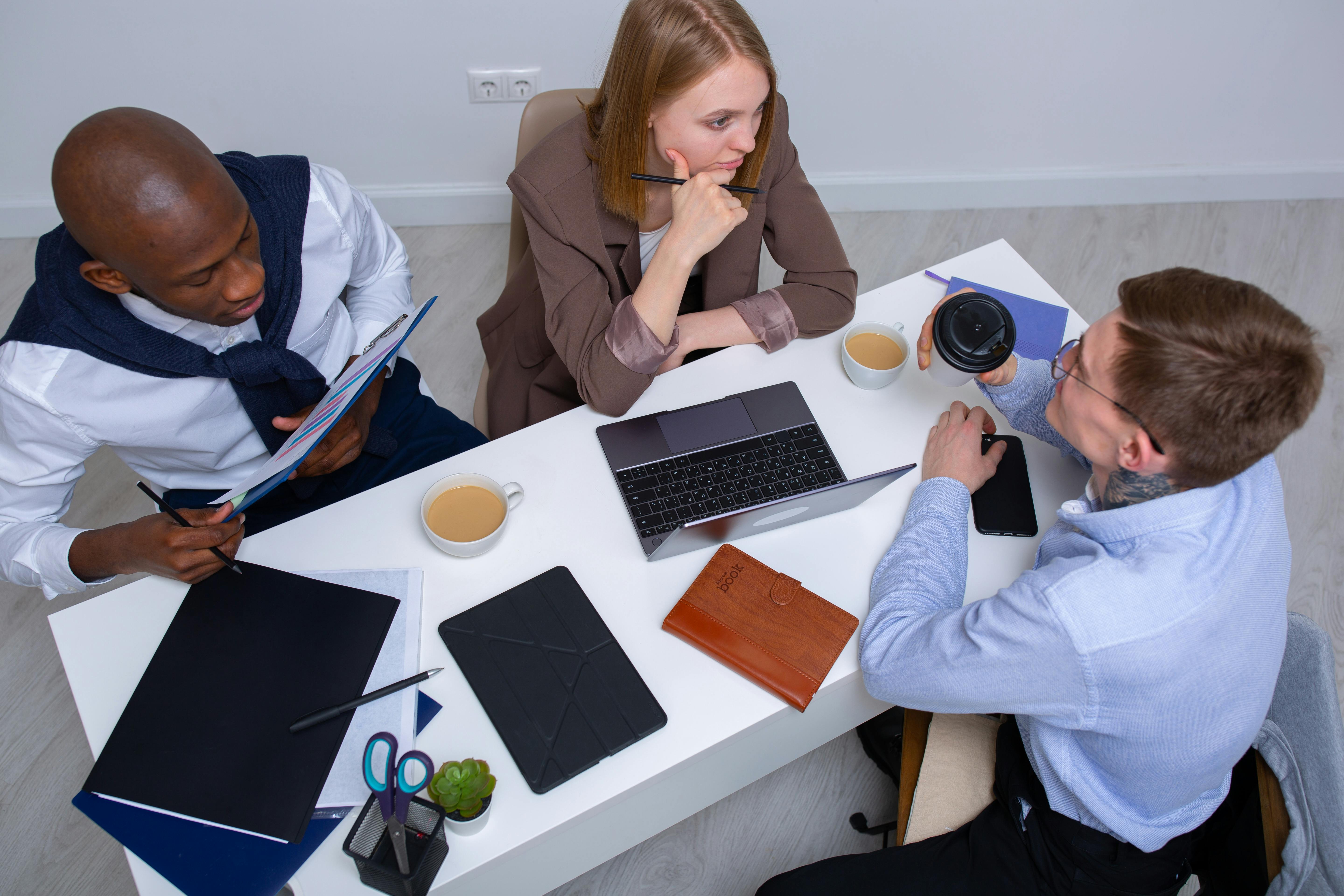 Three colleagues having a discussion around a white table with laptops, notebooks, coffee cups, and stationery.