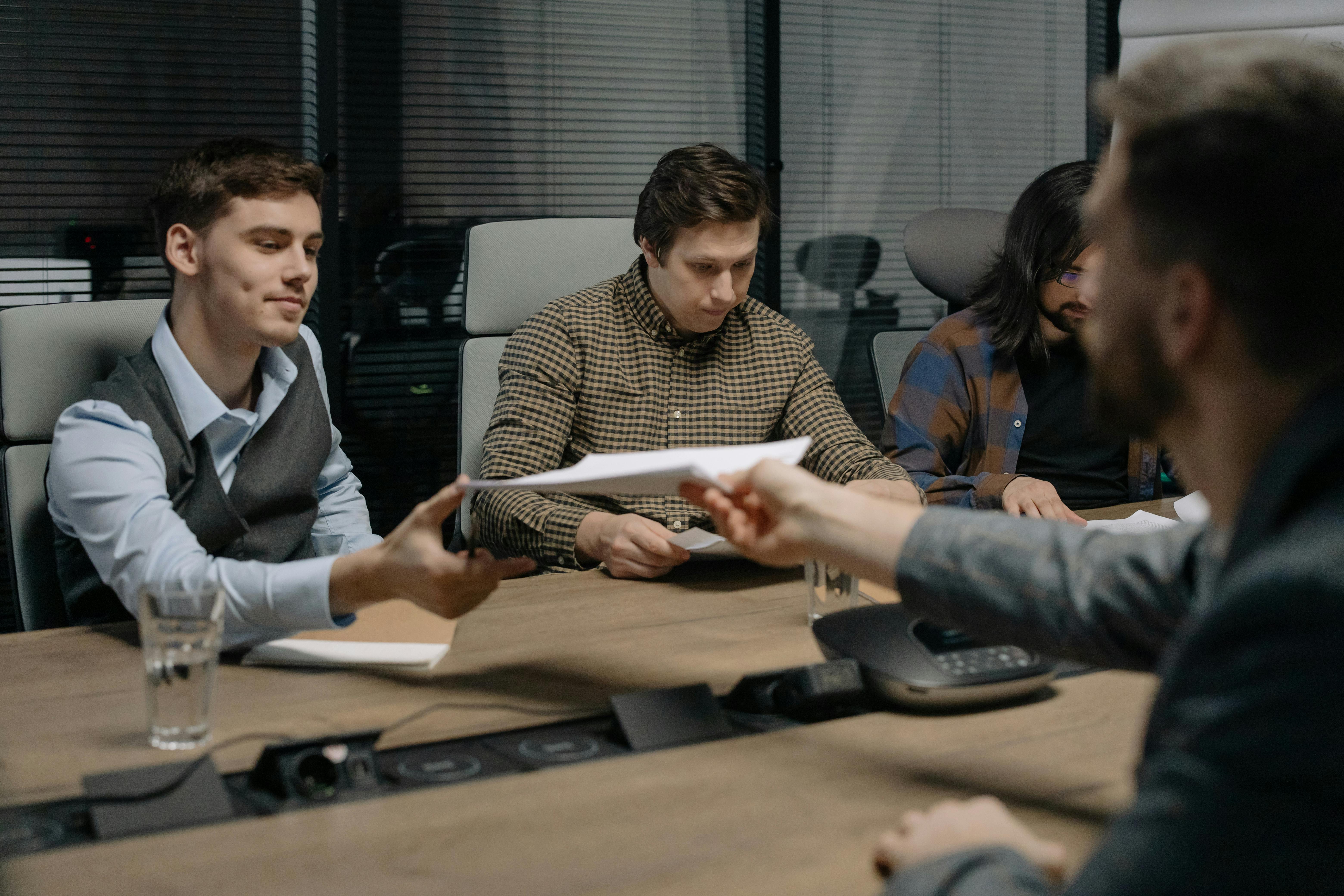 Four men sitting around a conference table, one man handing over papers to another in a boardroom setting.