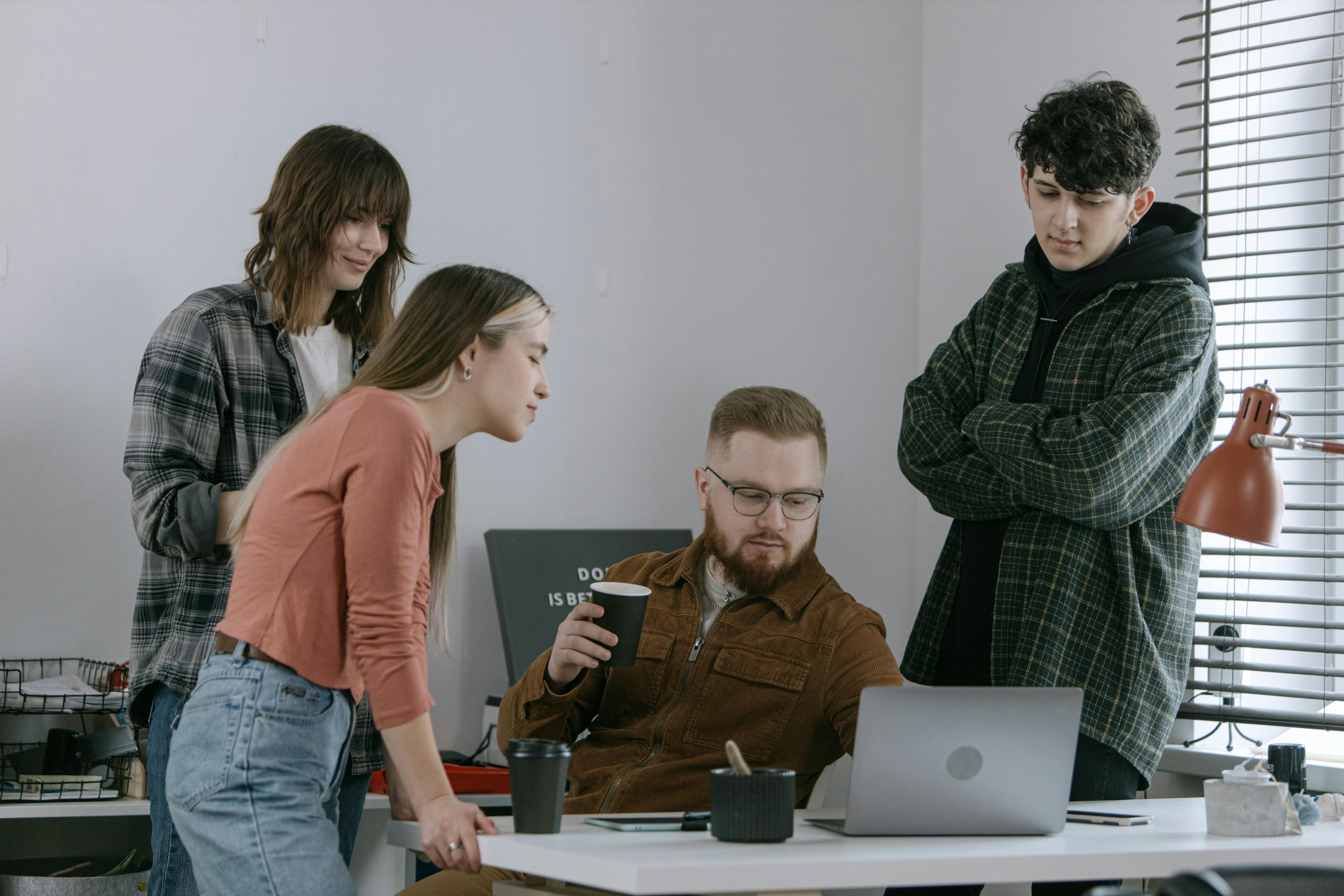 Four young adults gathered around a table, looking at a laptop, with two holding disposable coffee cups.