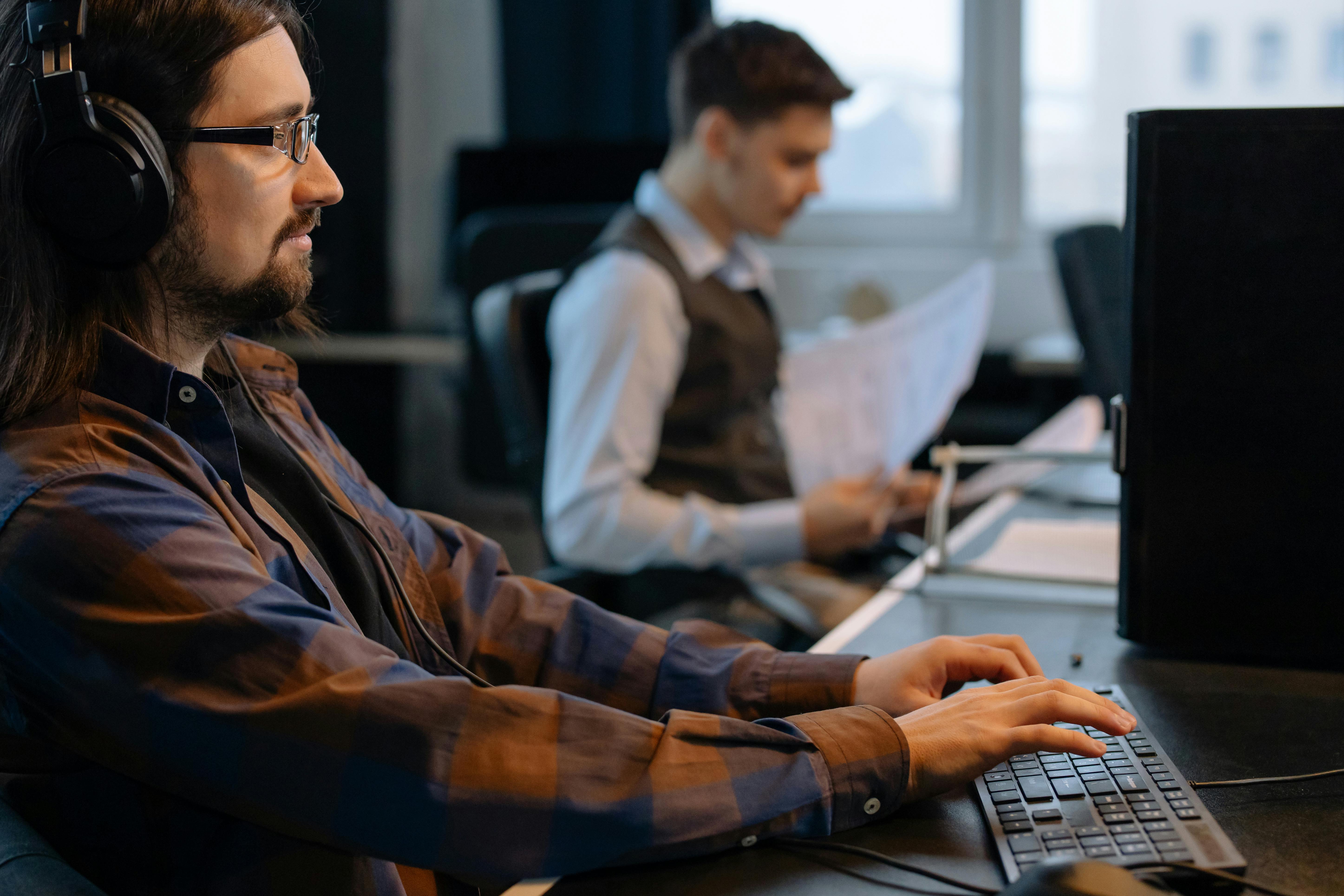 Two men working in an office; one wearing headphones types at a computer keyboard, while the other in a vest reads documents.