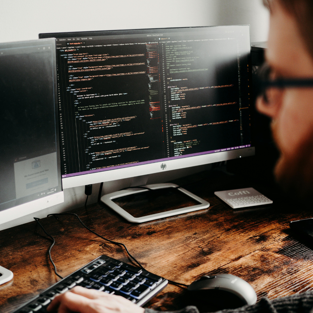 Close-up of a person coding on a dual-monitor setup with programming code displayed on one screen.