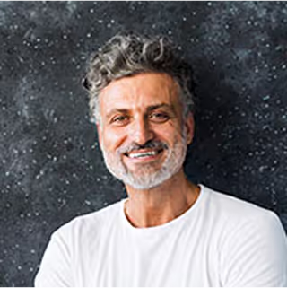 Smiling middle-aged man with curly salt-and-pepper hair and beard wearing a white shirt against a dark speckled background.