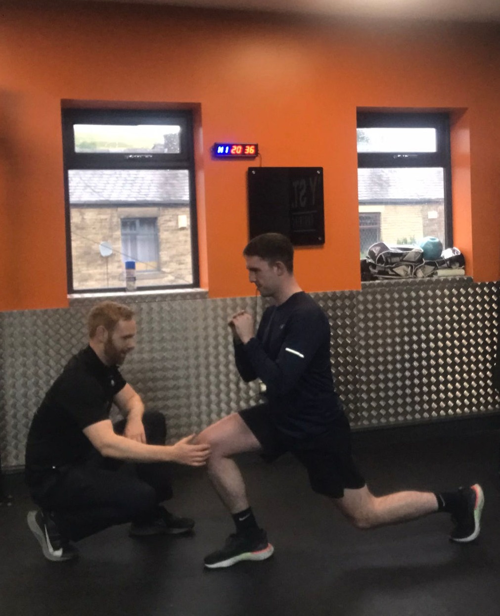 Trainer observing and supporting a man performing a lunge exercise in a gym with orange walls and two windows.