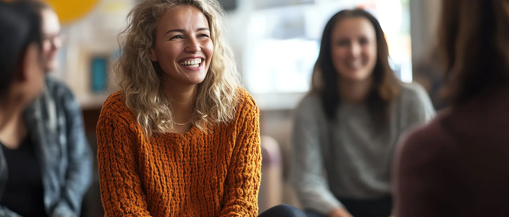 Smiling woman in an orange sweater engaged in conversation with three others in a cozy indoor setting.