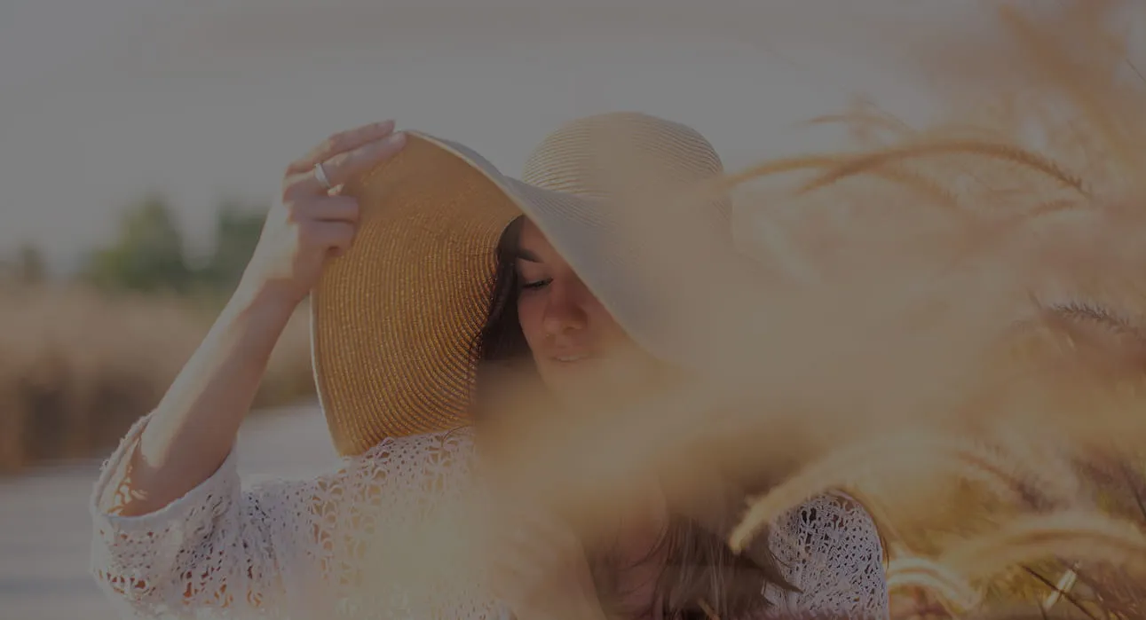 Woman wearing a large sunhat and white lace blouse, standing in a field with golden grass during sunset.