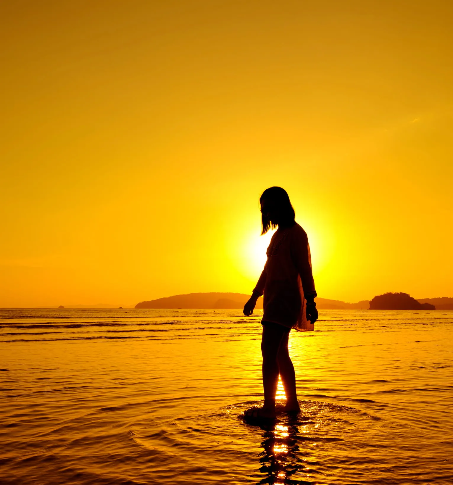 Silhouette of a person standing in shallow water during a golden sunset at the beach.