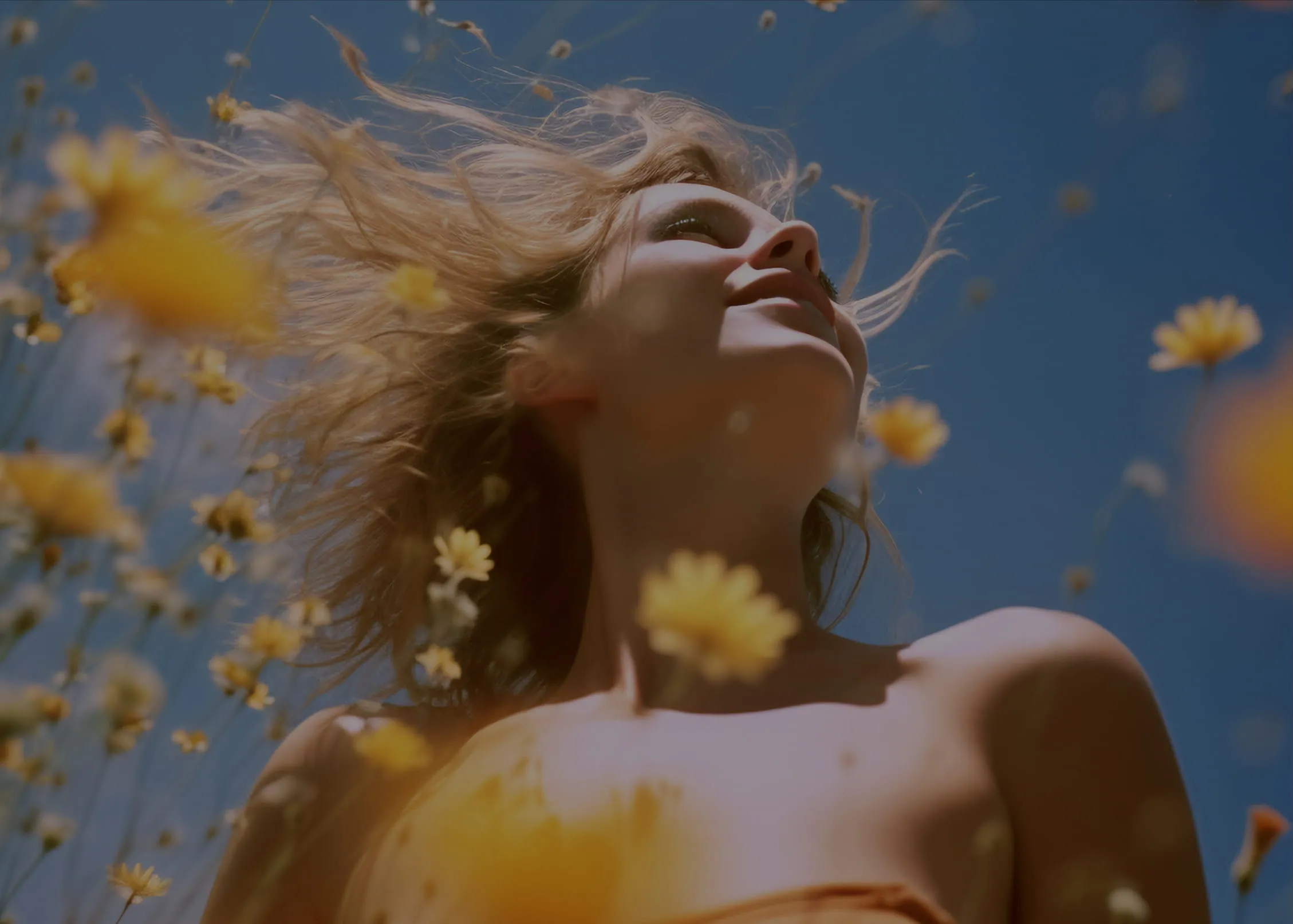 Young woman with flowing hair smiling among yellow wildflowers under a clear blue sky.