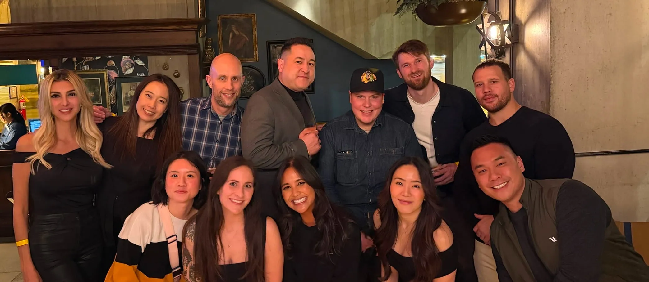Group of twelve diverse adults smiling and posing in a warmly lit indoor setting.