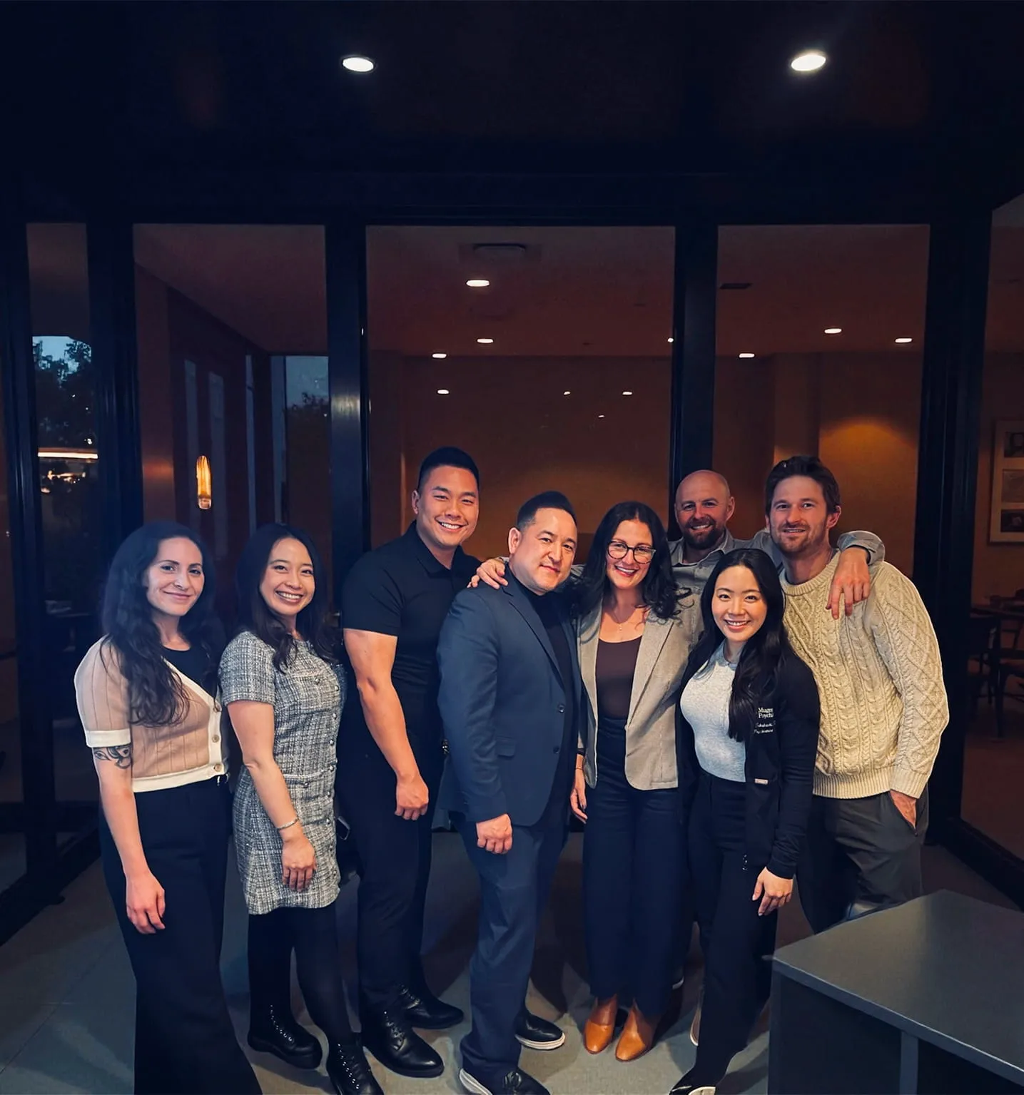 Eight smiling adults standing closely together in an indoor setting with glass doors behind them.