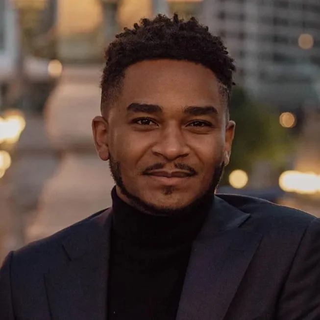 Portrait of a smiling man with short curly hair and a trimmed beard, wearing a dark turtleneck and suit jacket against an urban blurred background.