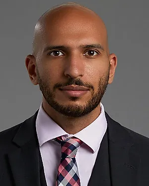 Portrait of a bald man with a beard wearing a black suit, light pink shirt, and a checkered tie against a gray background.